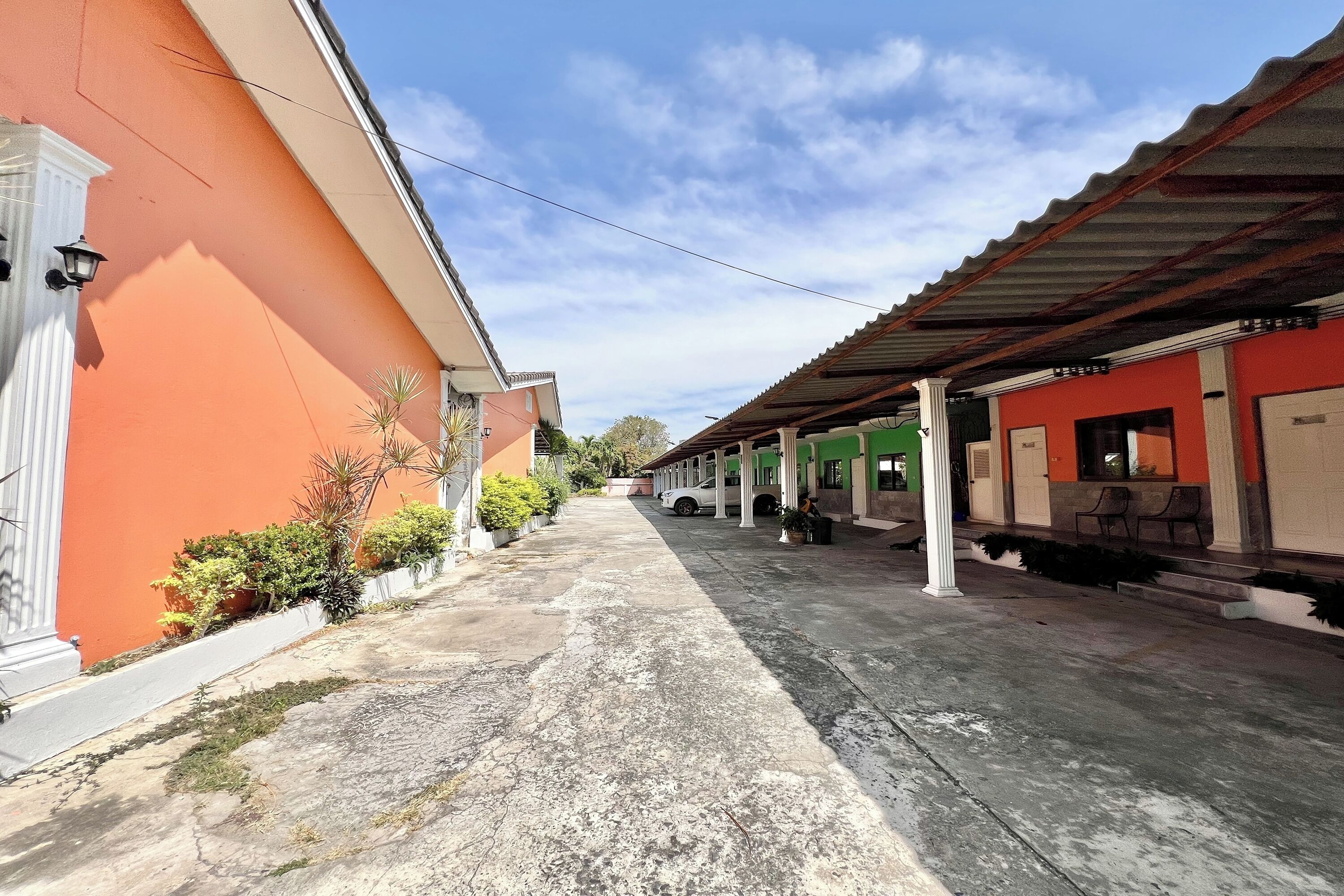 Un patio con suelo de concreto, un edificio de fachada naranja y una zona cubierta con paredes verdes y columnas blancas.