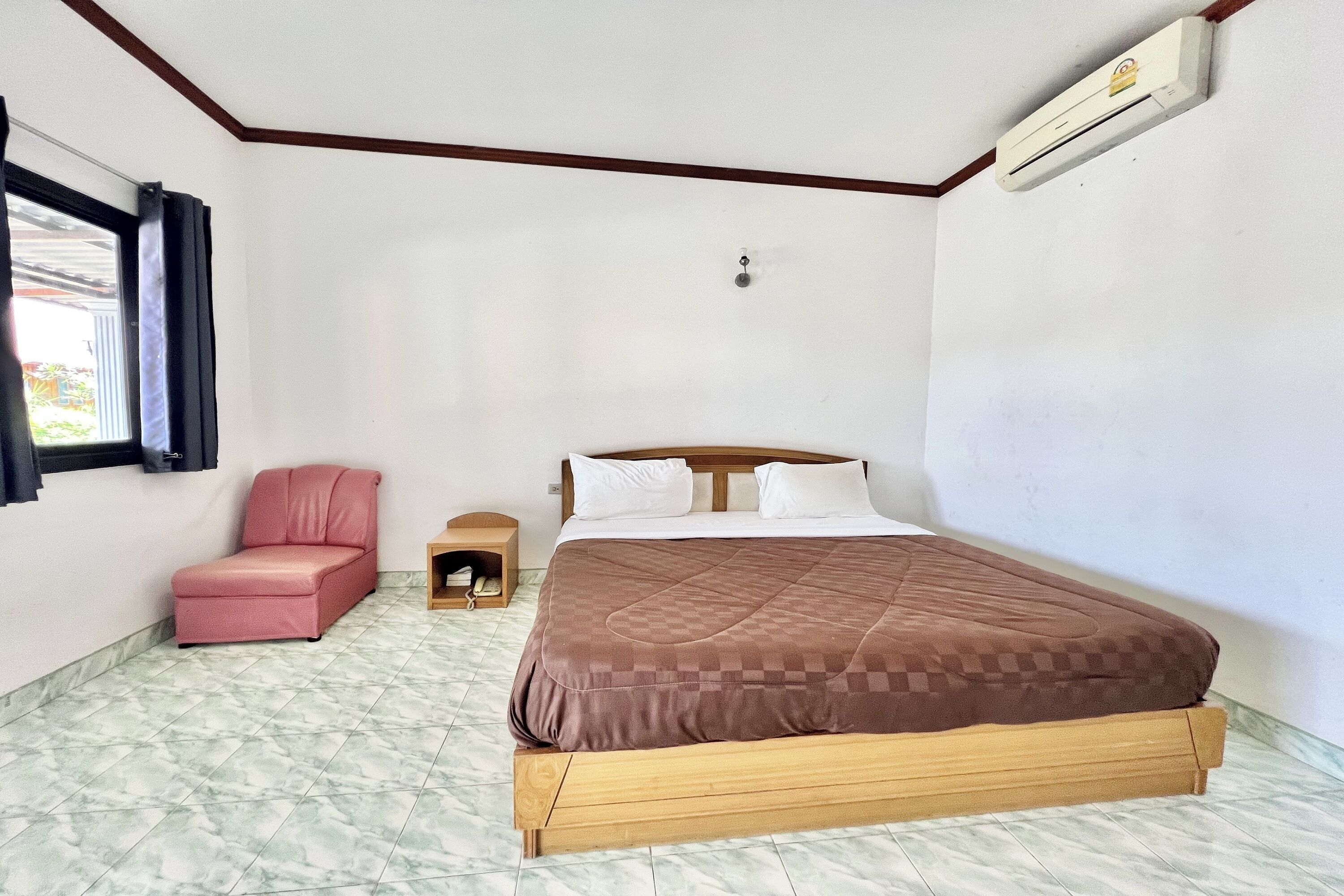 A bedroom with a bed, a pink chair, a small wooden cabinet, and an air conditioning unit.