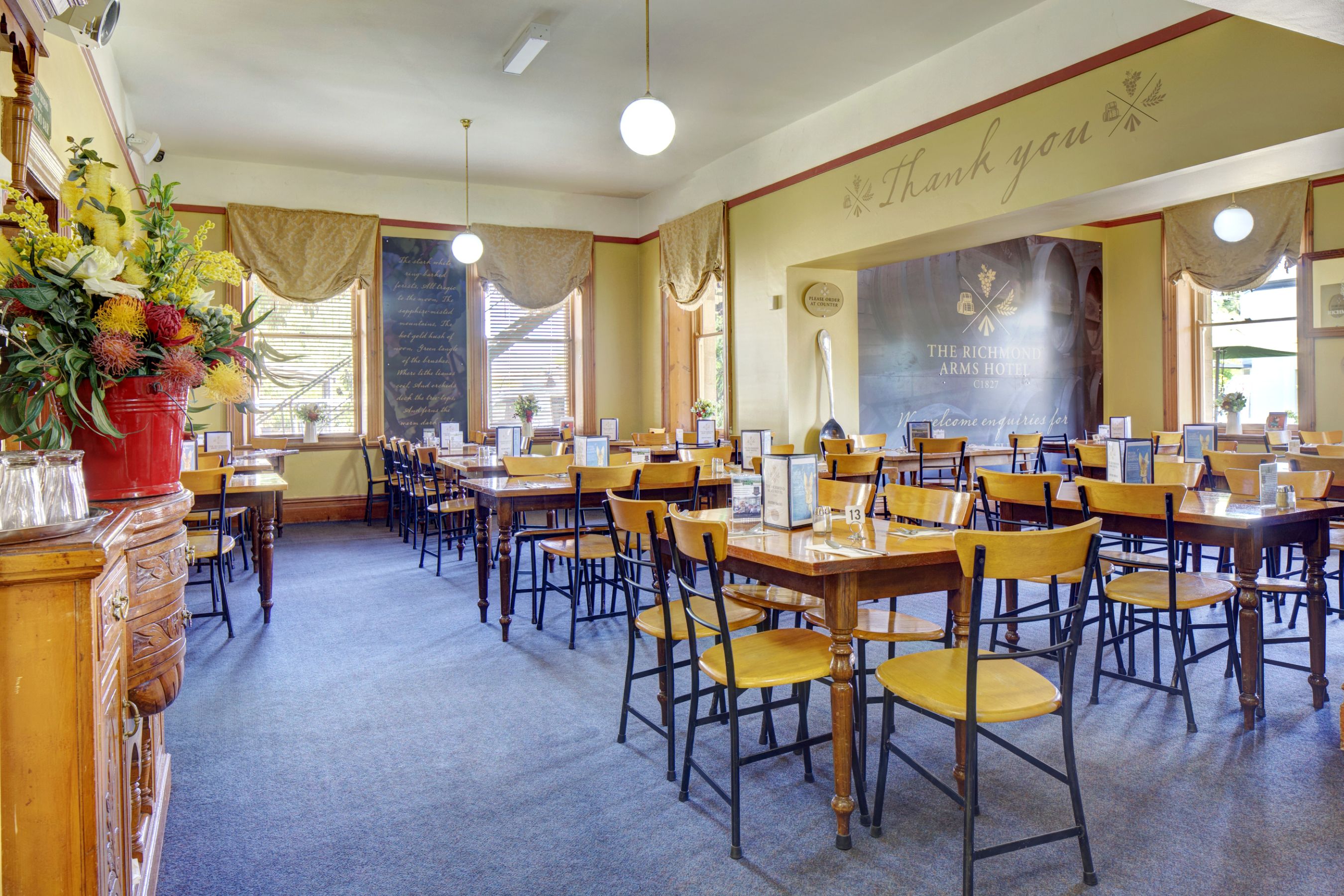 A hotel dining area with multiple tables and chairs, a large banner on the wall, and a flower arrangement on the left.