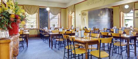 A hotel dining area with multiple tables and chairs, a large banner on the wall, and a flower arrangement on the left.