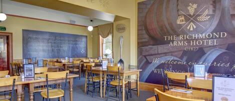 A dining area with multiple wooden tables and chairs, a chalkboard with writing, and a large mural of wine barrels.