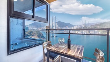 Un balcón con mesa, sillas y una botella de vino. Un ventanal grande ofrece vistas a un puente y al paisaje urbano.