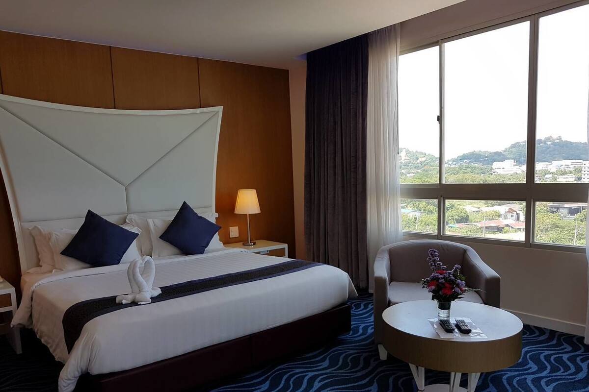 A hotel room with a large bed, a bedside table with a lamp, a chair, and a view of the city.