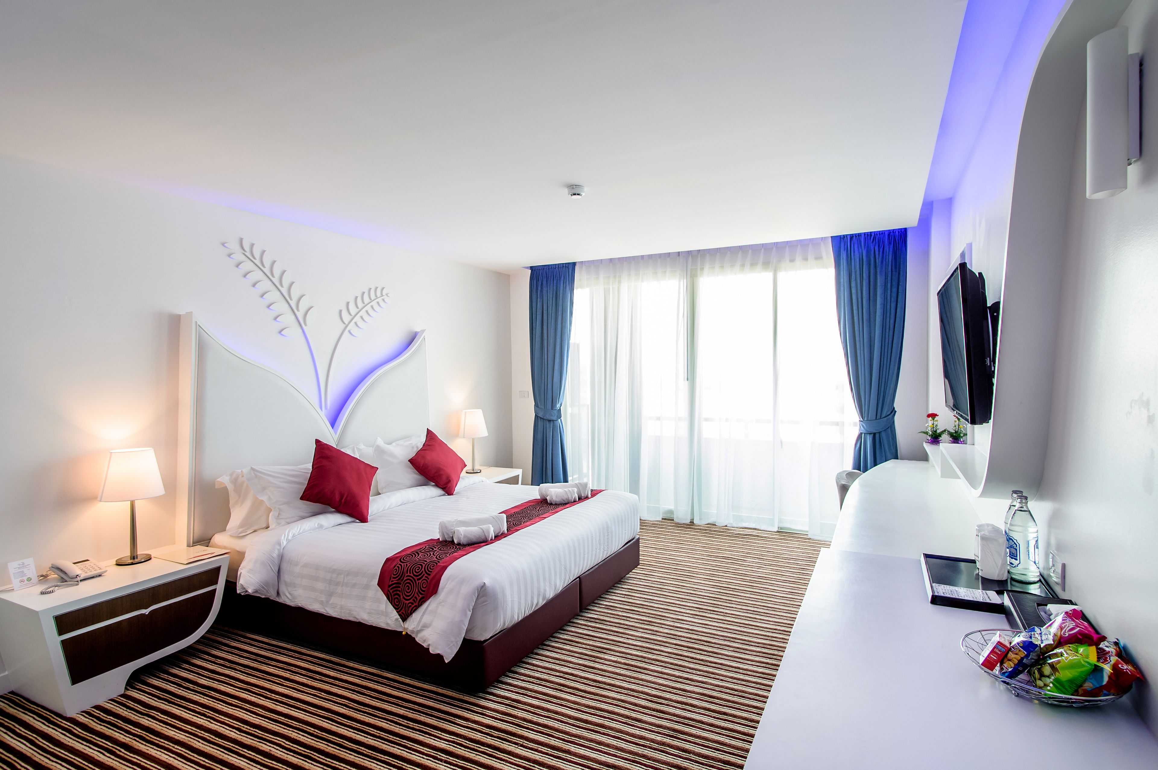 A hotel room with a bed, bedside table, lamp, and a desk with a computer and snacks.