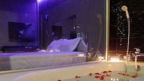 A modern bathroom with a bathtub filled with rose petals, a shower area, and a bed with a city view. - Heaven Inn (Si Racha)