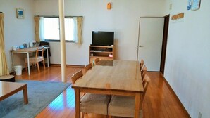 Una habitación con una mesa de comedor de madera, sillas, un televisor sobre un mueble bajo, un escritorio pequeño con una silla y una ventana con cortinas.