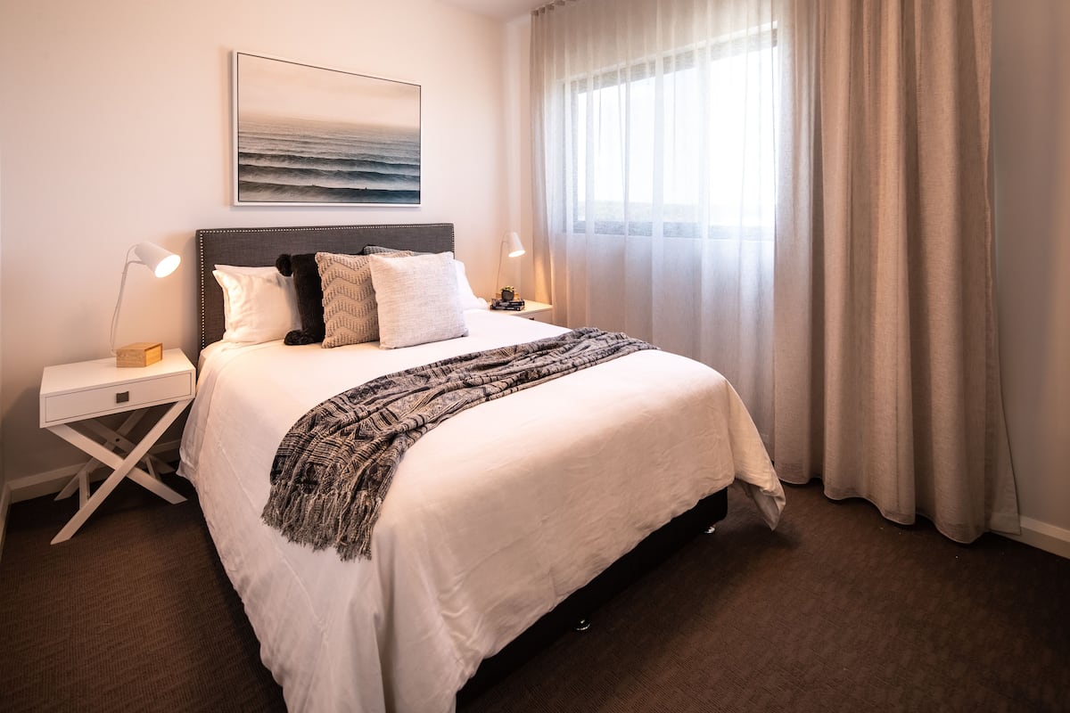 A neatly made bed with a headboard, a bedside table with a lamp, a framed picture on the wall, and a window with curtains.