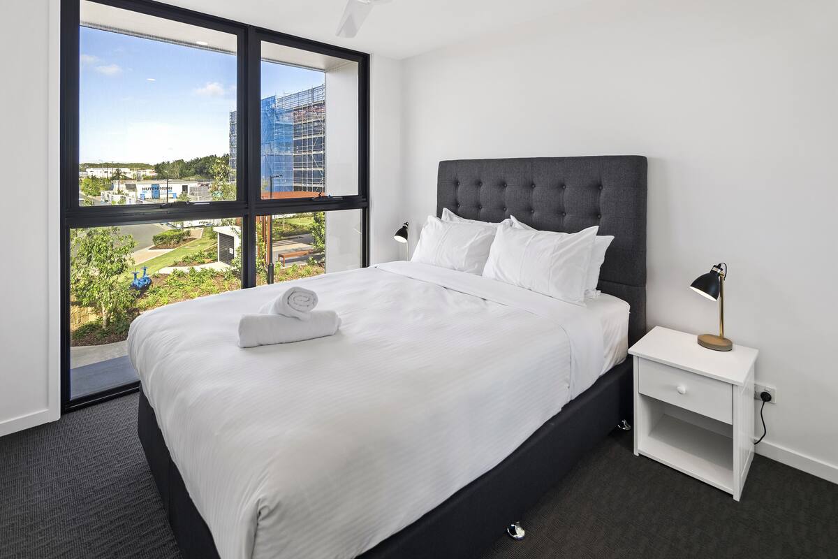 A hotel room with a large bed, a nightstand, a lamp, and a view of a construction site through a large window.