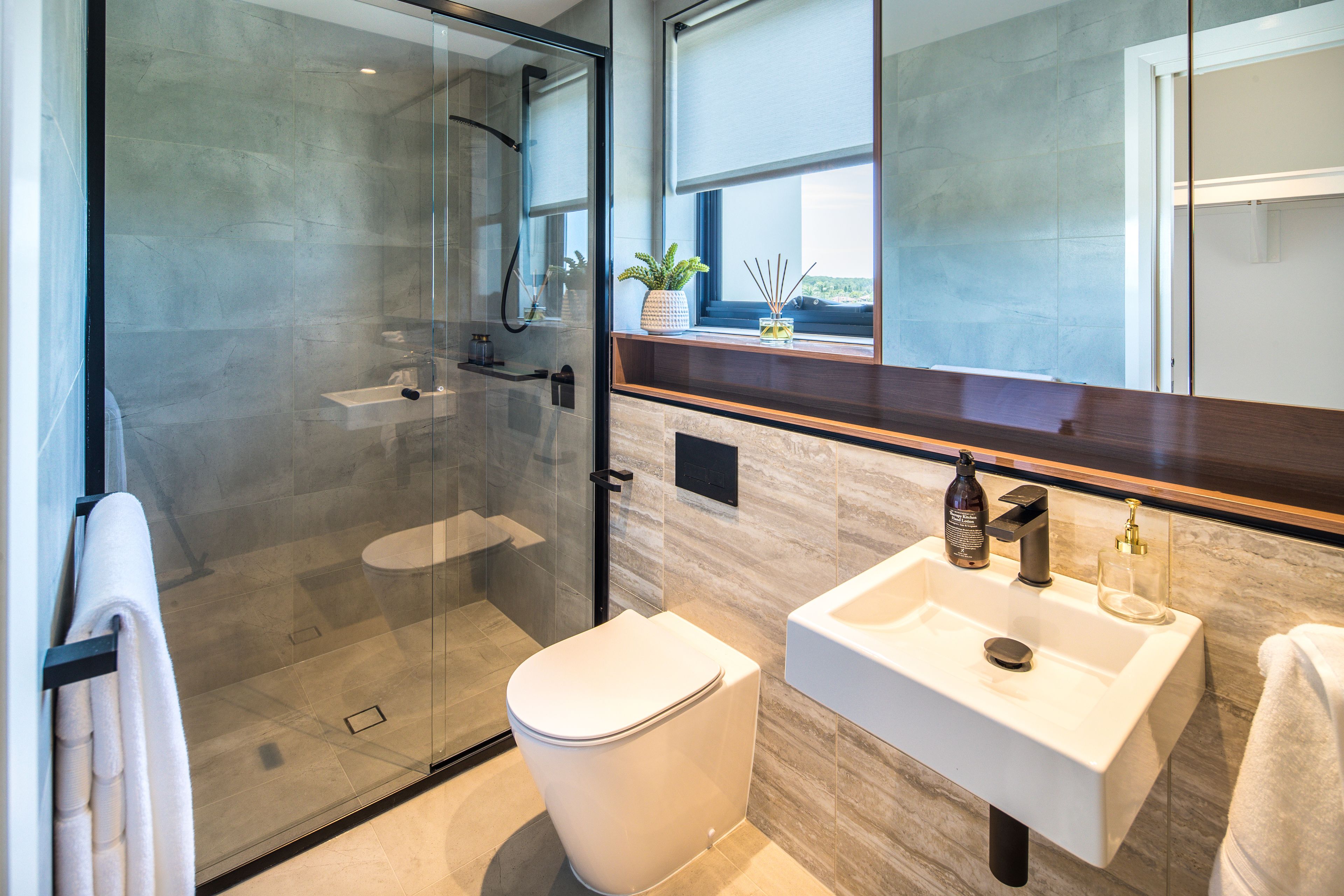 Une salle de bain moderne avec une douche à parois de verre, des toilettes blanches, un lavabo avec un miroir et une fenêtre ornée d’une plante posée sur le rebord.
