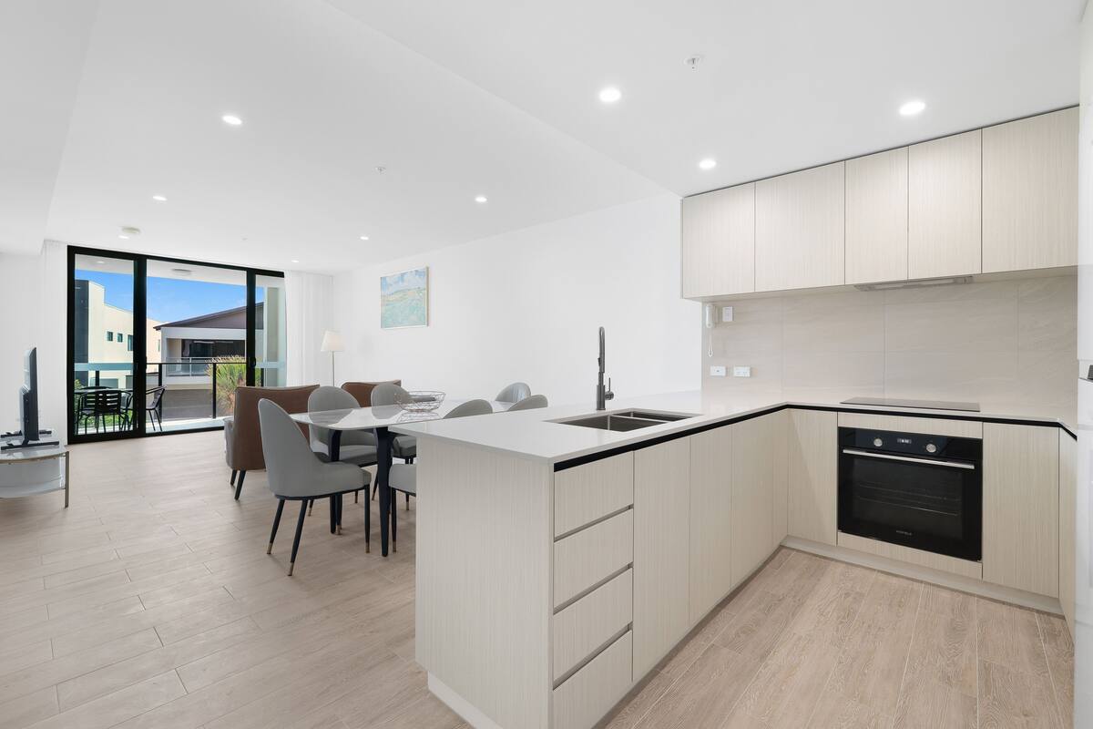 A modern kitchen with a built-in oven, a dining area with a table and chairs, and a large window offering a view of the outside.