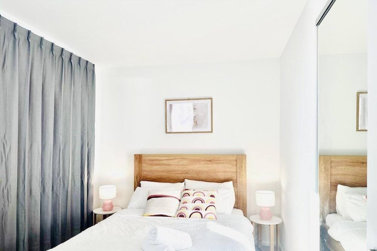 A hotel room with a wooden headboard, white bedding, a large window with grey curtains, two bedside tables with lamps, and a framed picture on the wall.