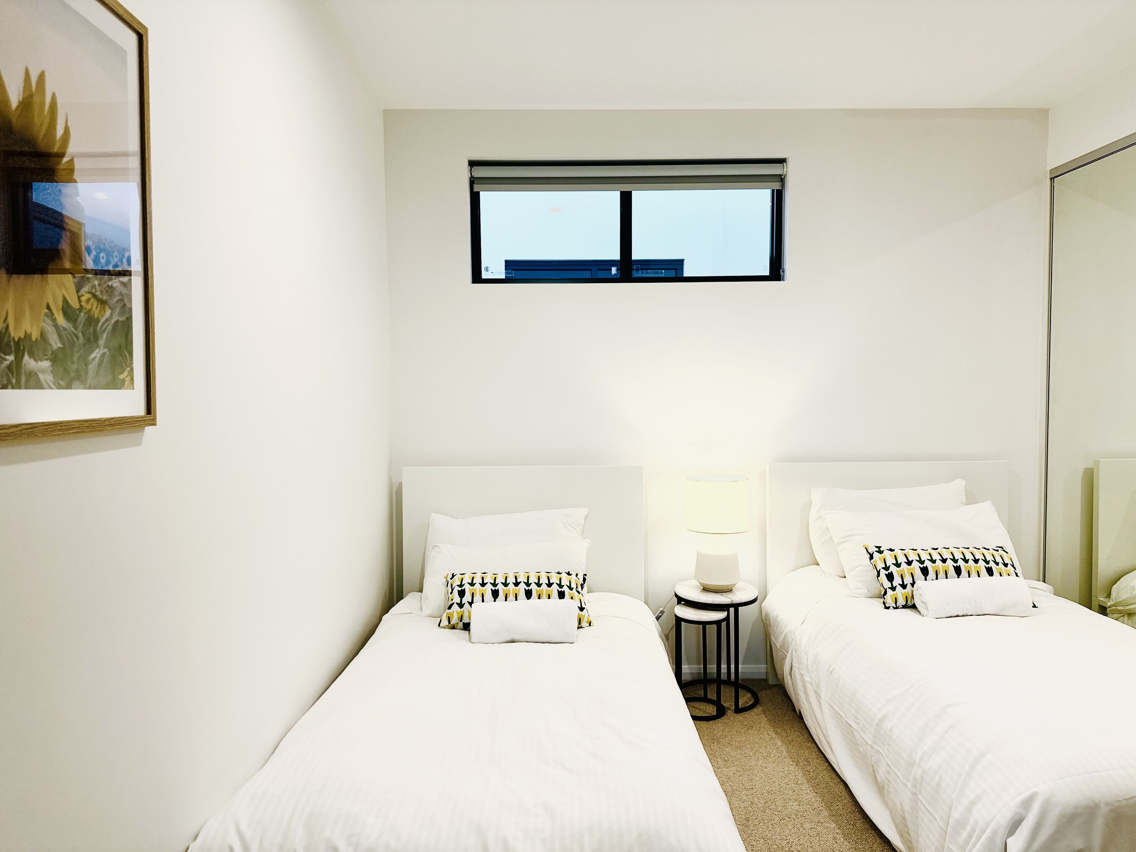 A hotel room with two beds, a small table with a lamp, and a window with a view of the sky.