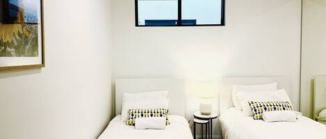 A hotel room with two beds, a small table with a lamp, and a window with a view of the sky.
