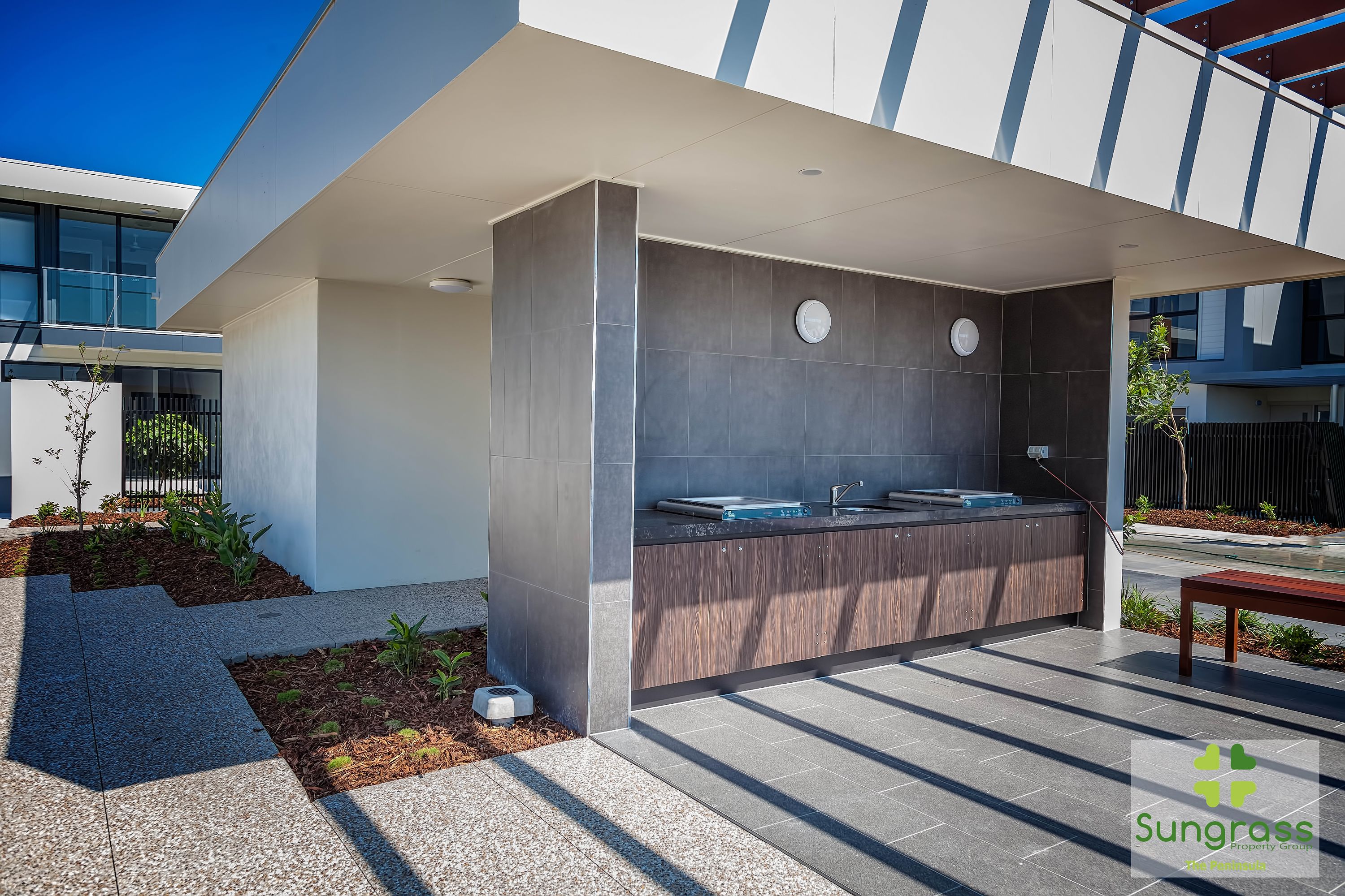Un'area cucina esterna moderna con un bancone integrato e due lavandini, circondata da un patio con una panchina e un piccolo giardino.