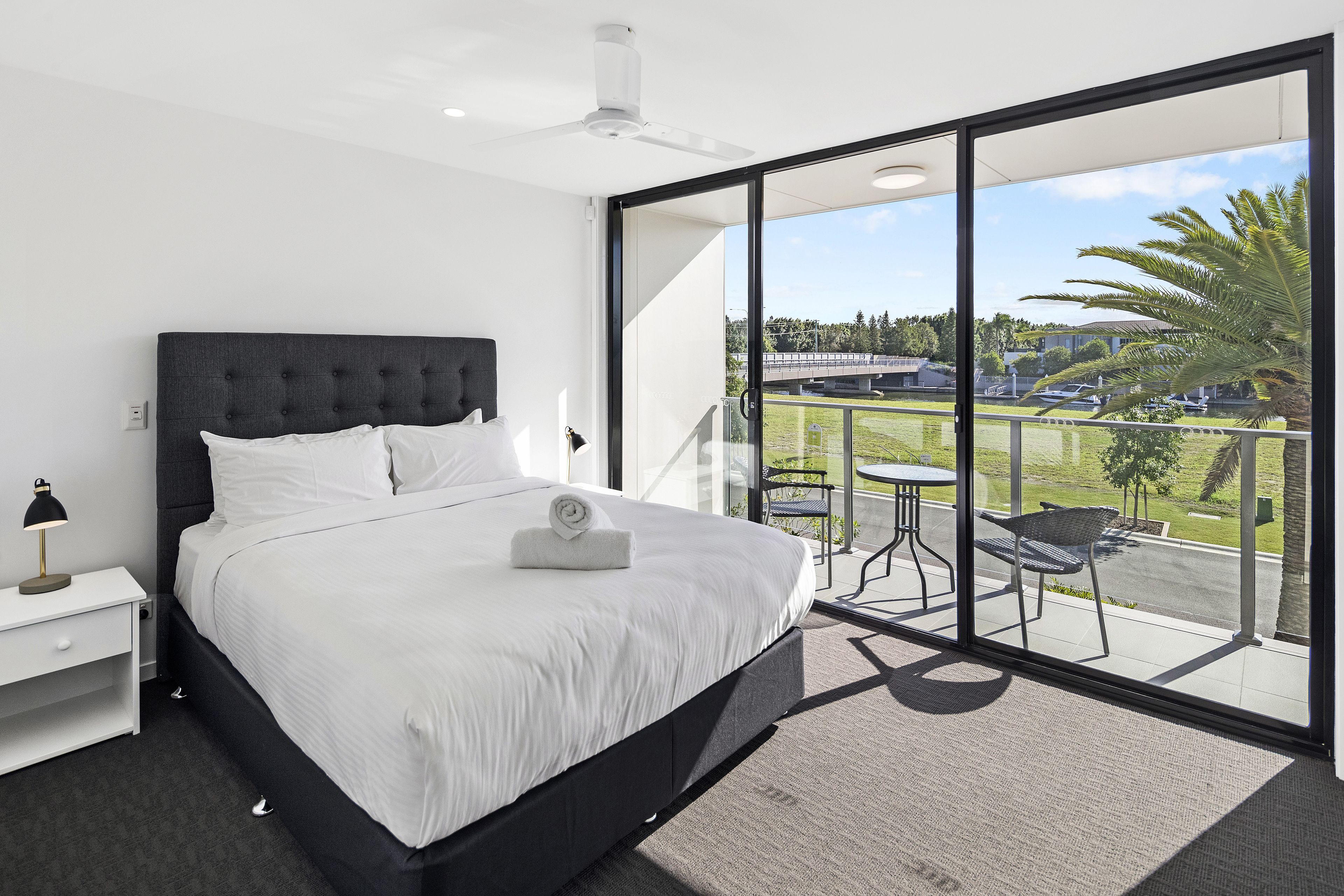 A modern bedroom with a large bed, a nightstand, a desk, and a balcony with a view of the outdoors.