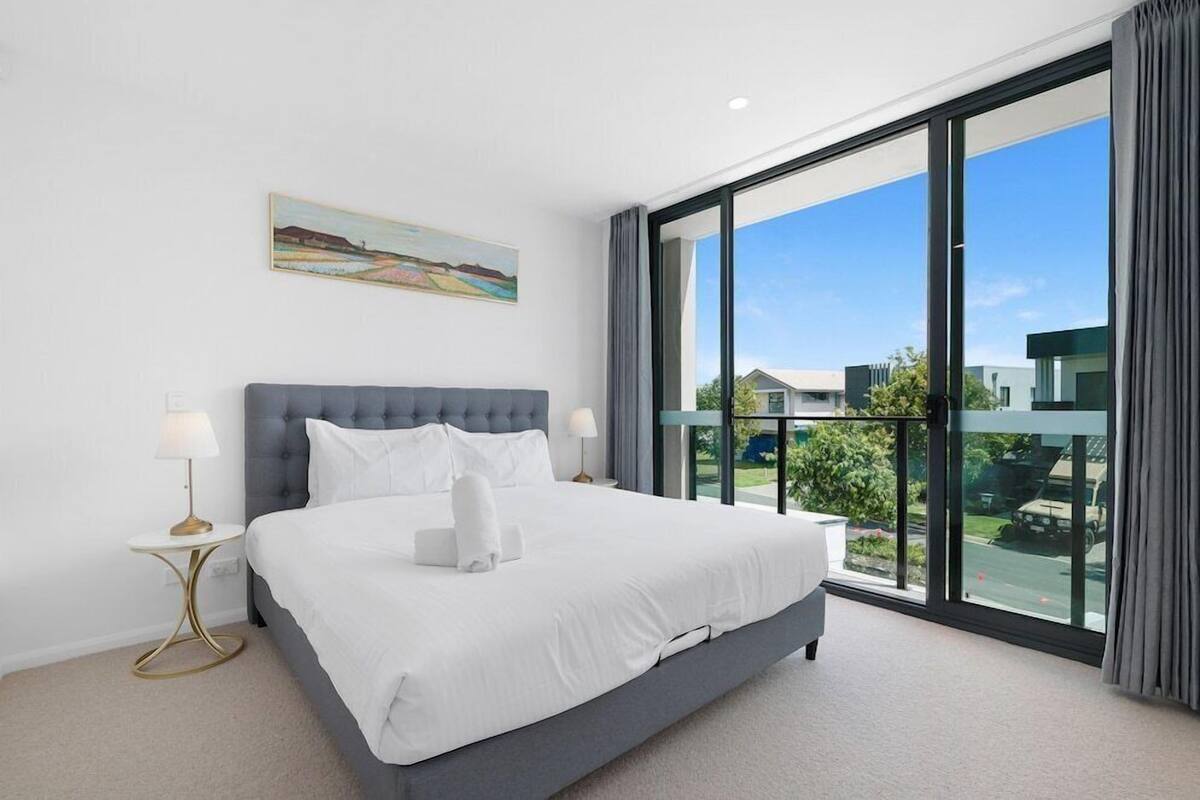 A modern hotel room with a large bed, two bedside tables, a painting on the wall, and a balcony with a view.