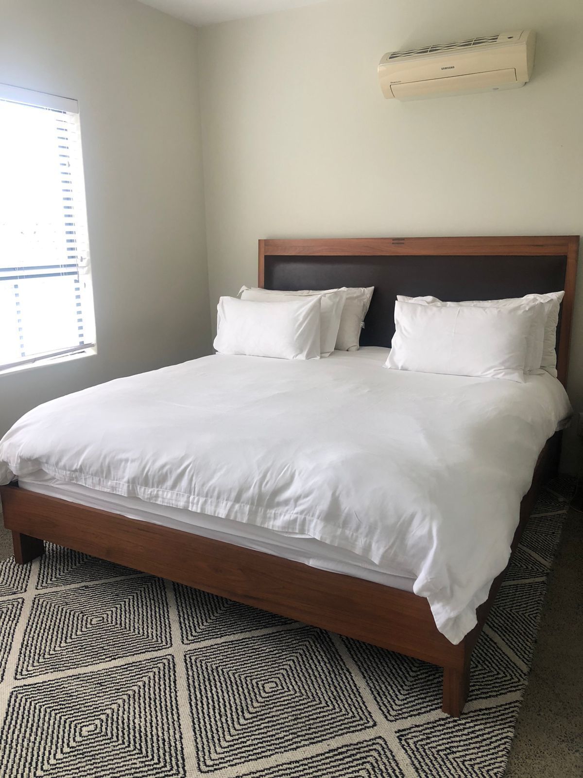 A neatly made bed with white linens in a room with a patterned rug, a window with blinds, and a wall-mounted air conditioning unit.