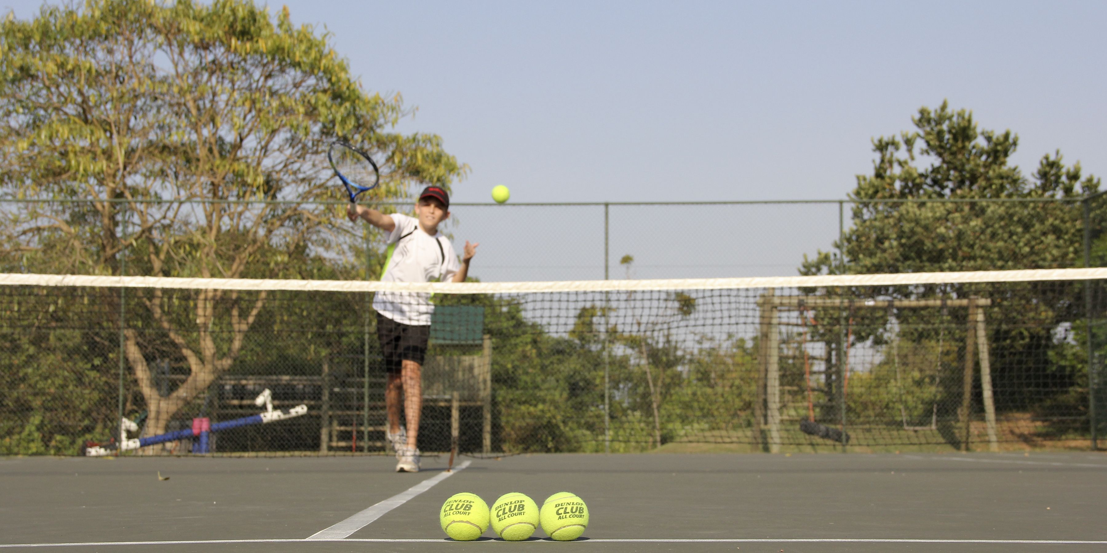 Ein Tennisplatz mit Netz, ein Spieler im Spiel und Tennisbälle am Boden.