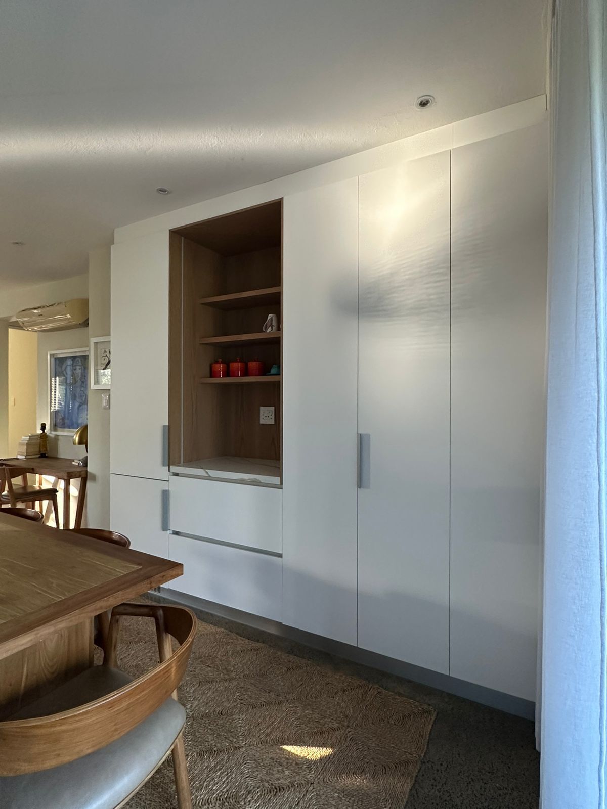 A modern kitchen with white cabinets, a built-in wooden shelf, and a dining area with a wooden table and chairs.
