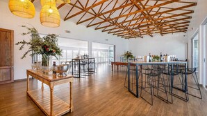 Una habitación amplia con techo alto, vigas de madera y ventanas grandes. Cuenta con una mesa de comedor con sillas y una zona de bar con botellas.