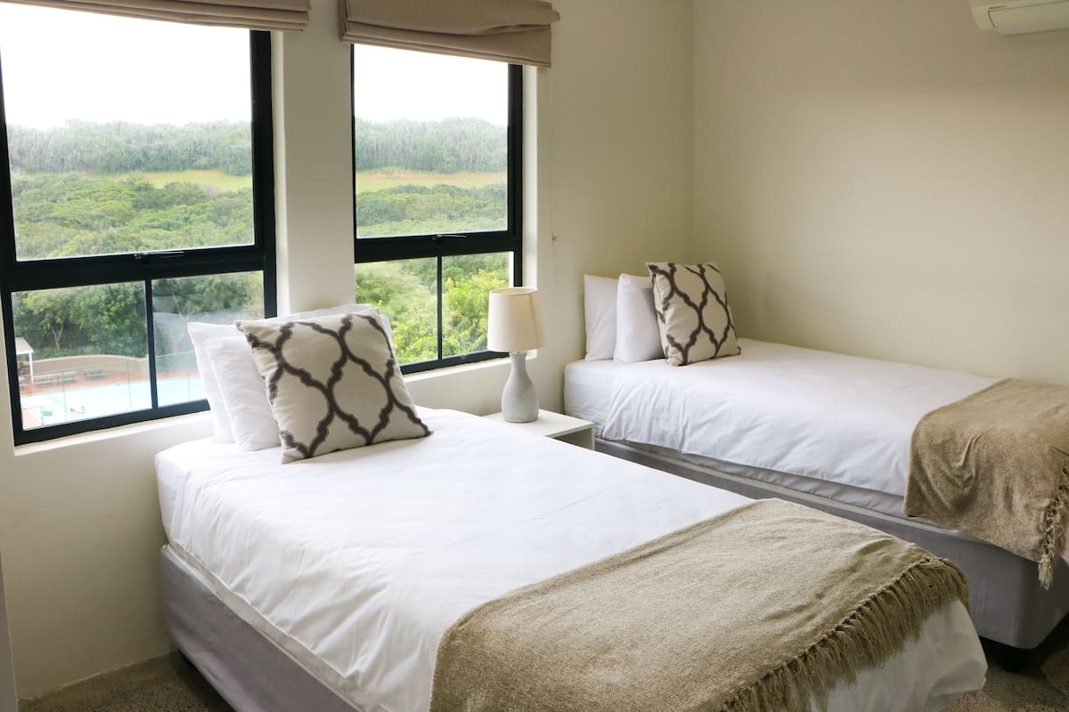 Une chambre avec deux lits, une table de chevet, une lampe et une vue sur une forêt, visible à travers de grandes fenêtres.