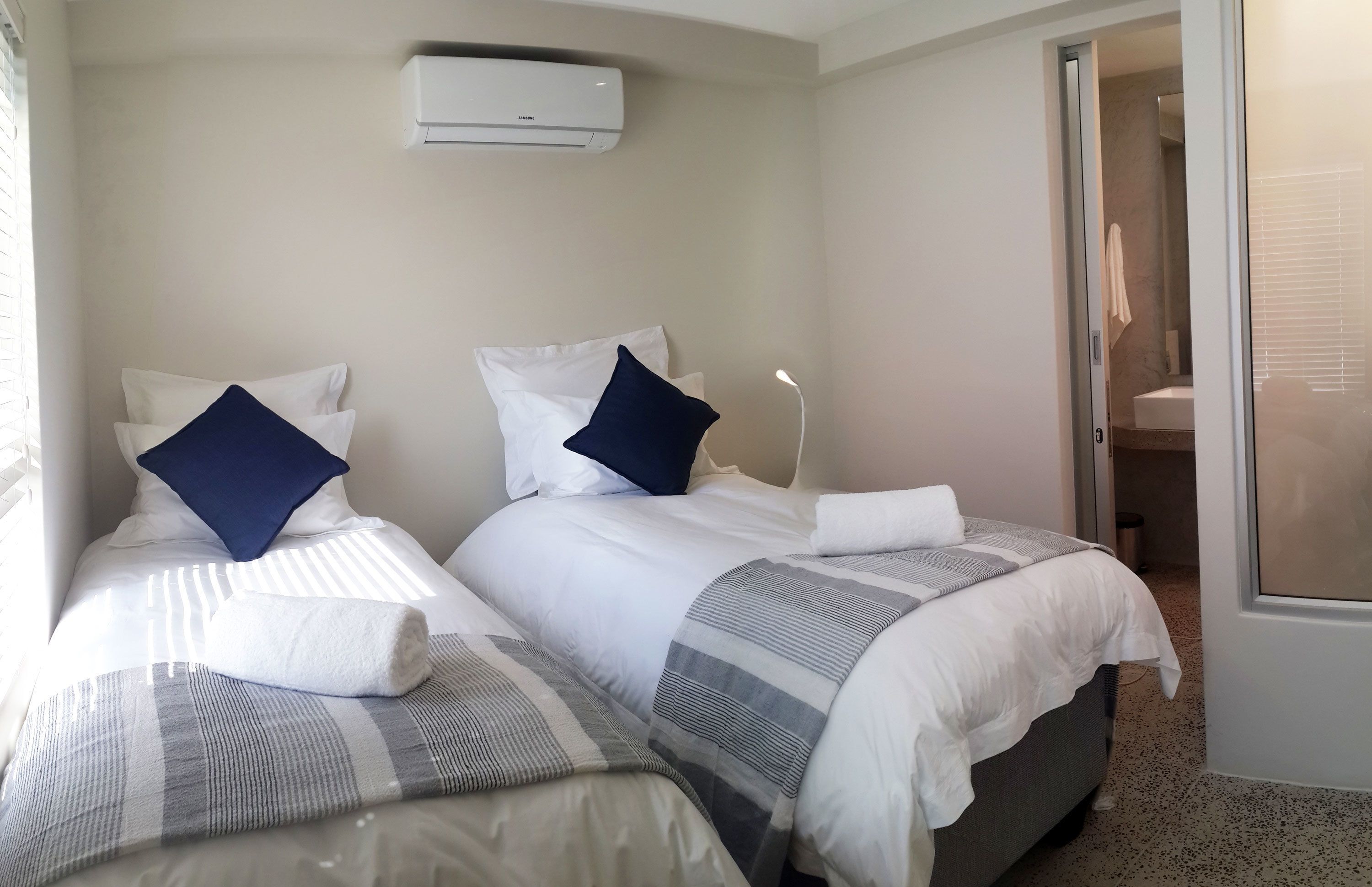 A hotel room with two beds, a window, an air conditioning unit, and a bathroom visible through an open door.