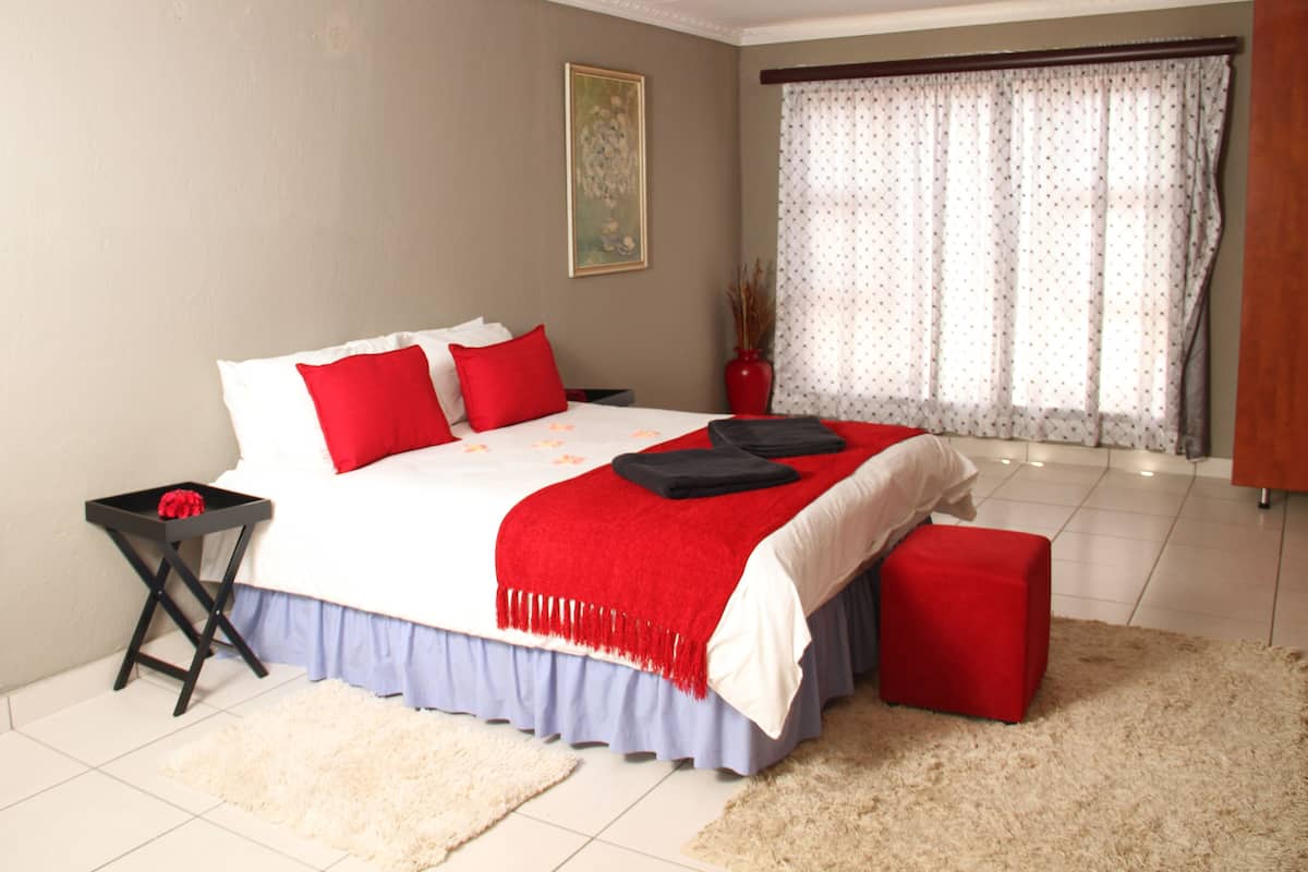 Une chambre à coucher avec un lit, une table de chevet, un banc rouge, un vase contenant des plantes séchées et une fenêtre avec des rideaux.