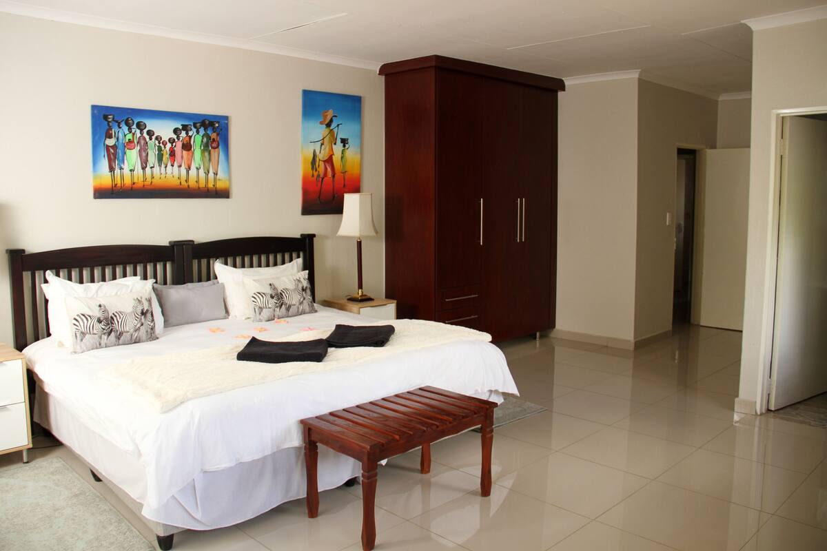 Quarto de hotel com uma cama grande, um banco de madeira, um guarda-roupa e duas pinturas na parede.