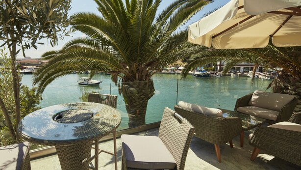 A terrace with wicker furniture, a glass table, and a view of a marina with boats. - Kubana Hotel (Cesme)