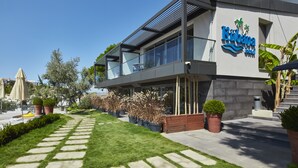 A modern hotel with a glass facade, outdoor seating, and a well-maintained garden. - Kubana Hotel (Cesme)