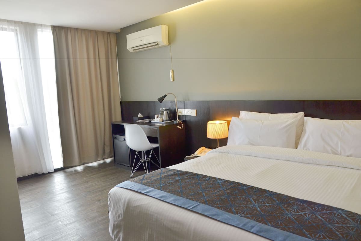 A hotel room with a bed, desk, chair, and an air conditioning unit.
