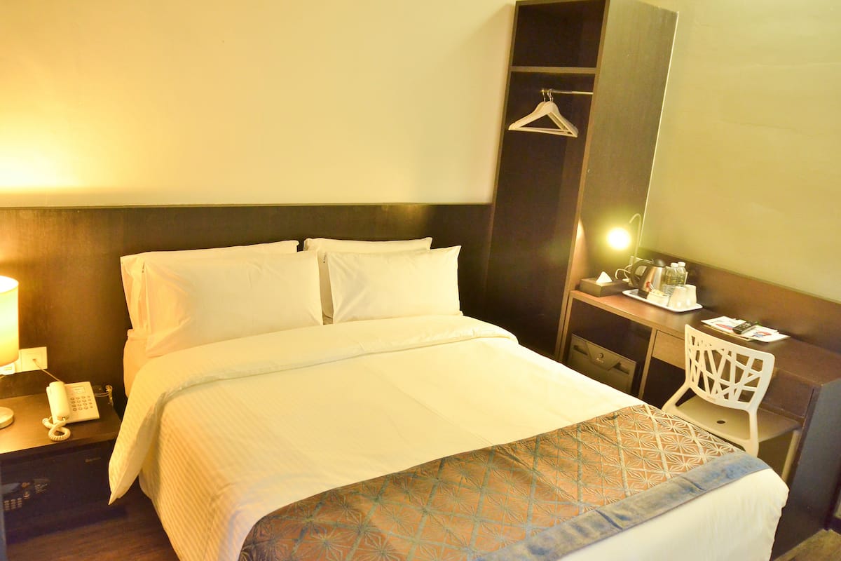 A hotel room with a large bed, a bedside table with a lamp, a small desk with a chair, and a mirror.