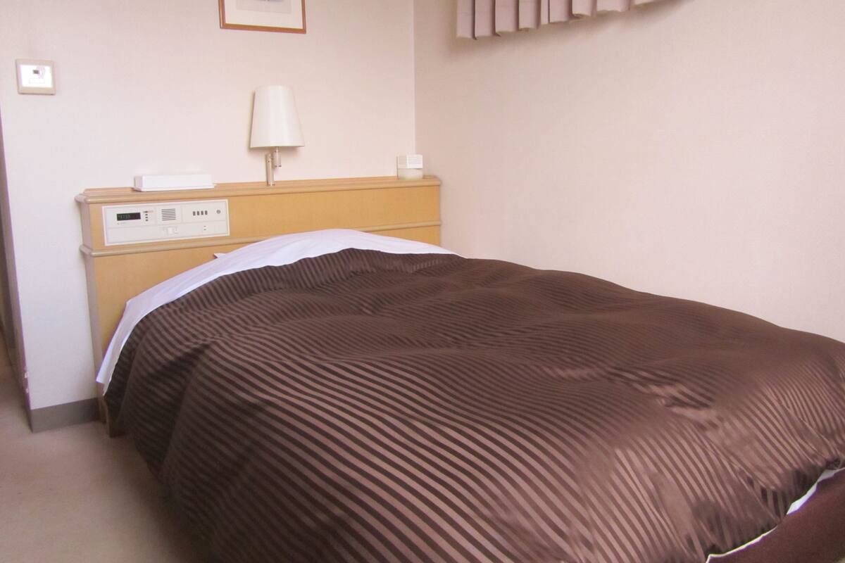 A hotel room with a bed, a bedside table, a lamp, and a thermostat.