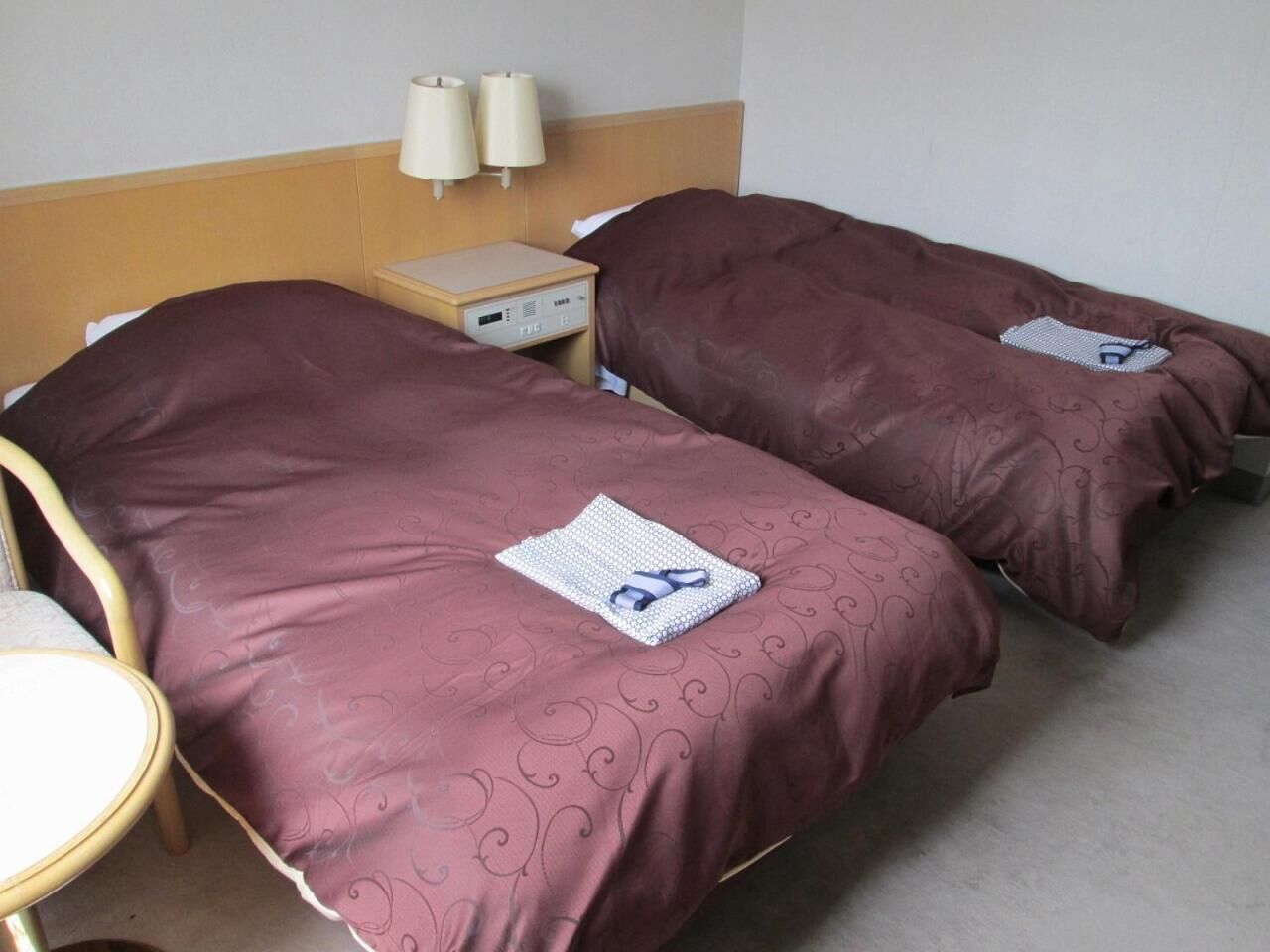 A hotel room with two beds, each with a brown bedspread and a folded towel with a blue and white pattern. There is a wooden nightstand with a lamp between the beds, and a chair with a white cushion.
