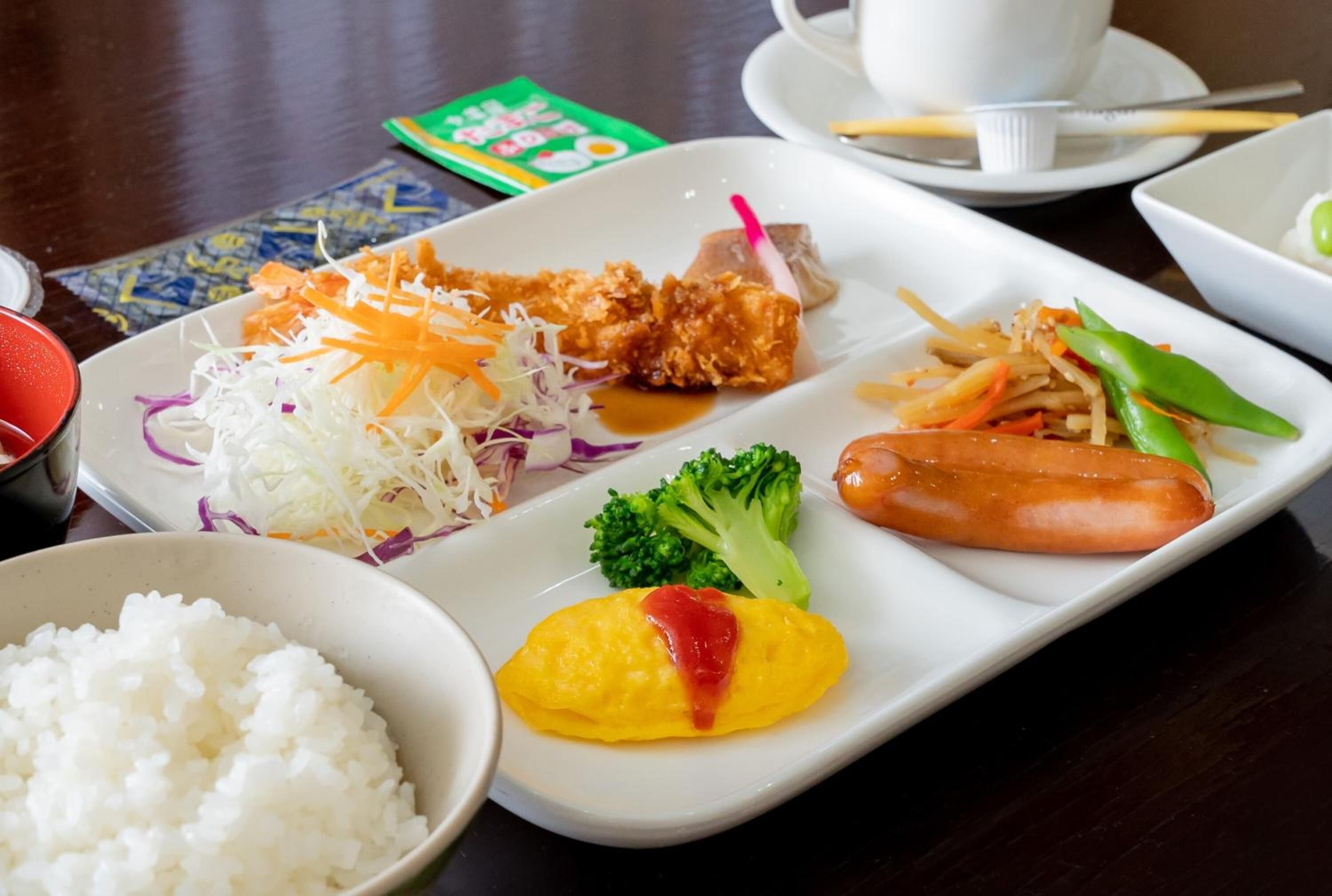 A meal with rice, a fried item, a sausage, a vegetable omelet, and a side of shredded cabbage and carrots.
