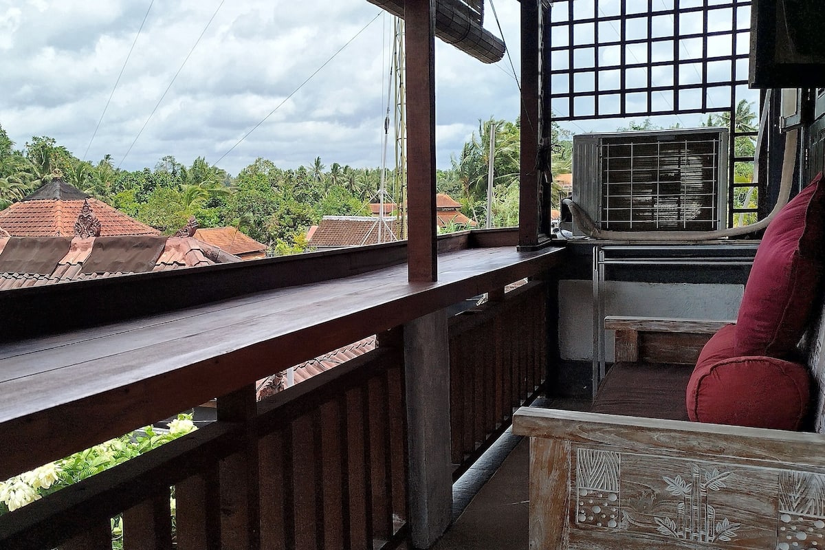 A balcony with a wooden railing, a red cushion, and a view of buildings with red roofs and palm trees.