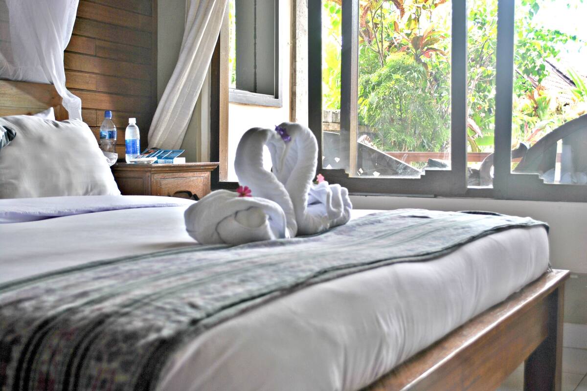 A bed with a canopy, a nightstand with a water bottle, and a window with a view of greenery.