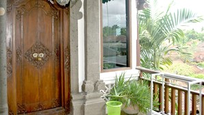 A wooden door with intricate carvings, a stone pillar, a balcony with a railing, and a potted plant. - Rumah Roda (Ubud)