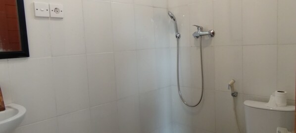 A bathroom with white tiles, a shower with a handheld showerhead, and a toilet with a paper towel holder. - Rumah Roda (Ubud)