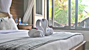 A neatly made bed with a white comforter and a floral-patterned blanket. There are two swans made of towels on the bed, and a bottle of water and a magazine on the nightstand beside the bed. - Rumah Roda (Ubud)