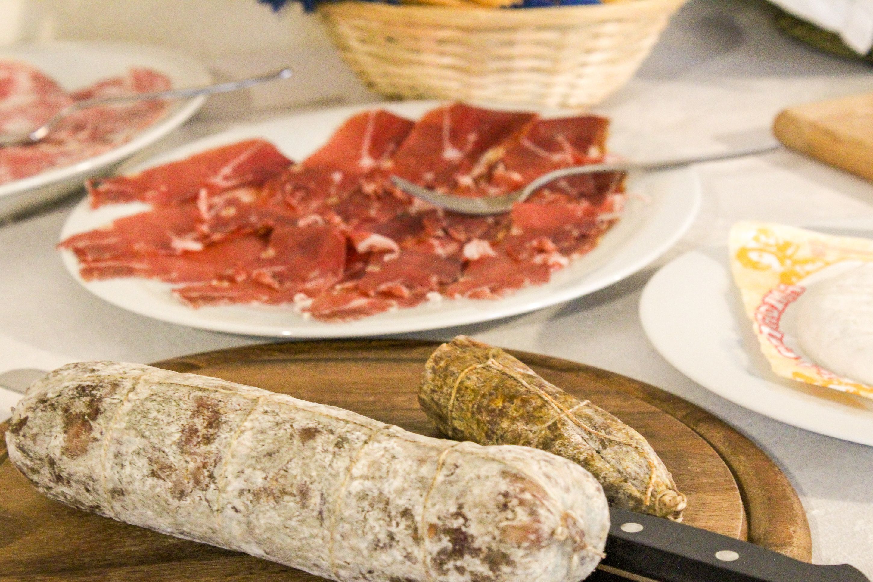 Un tavolo con diversi cibi tra cui affettati, una grossa salsiccia e un involucro, forse formaggio o pasta.