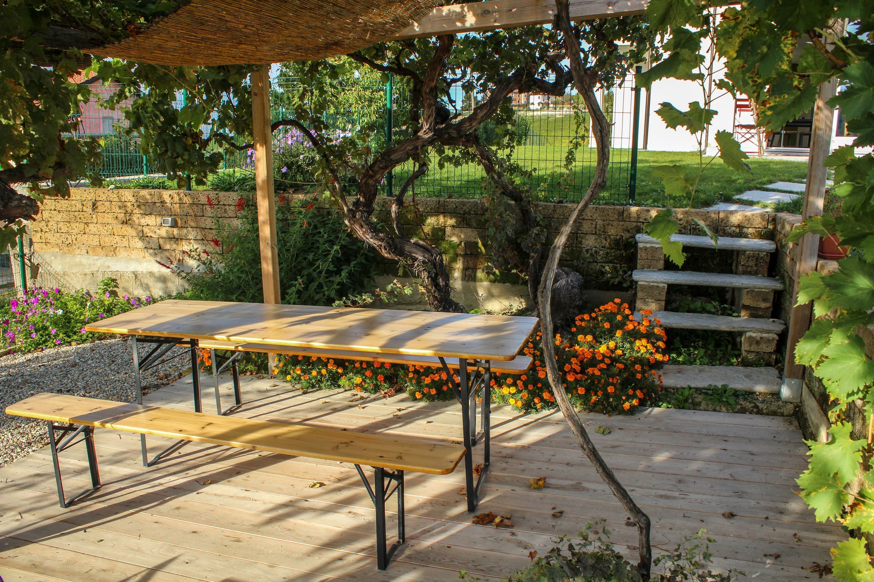 A wooden picnic table with benches under a pergola, surrounded by a garden with flowers and a stone wall.