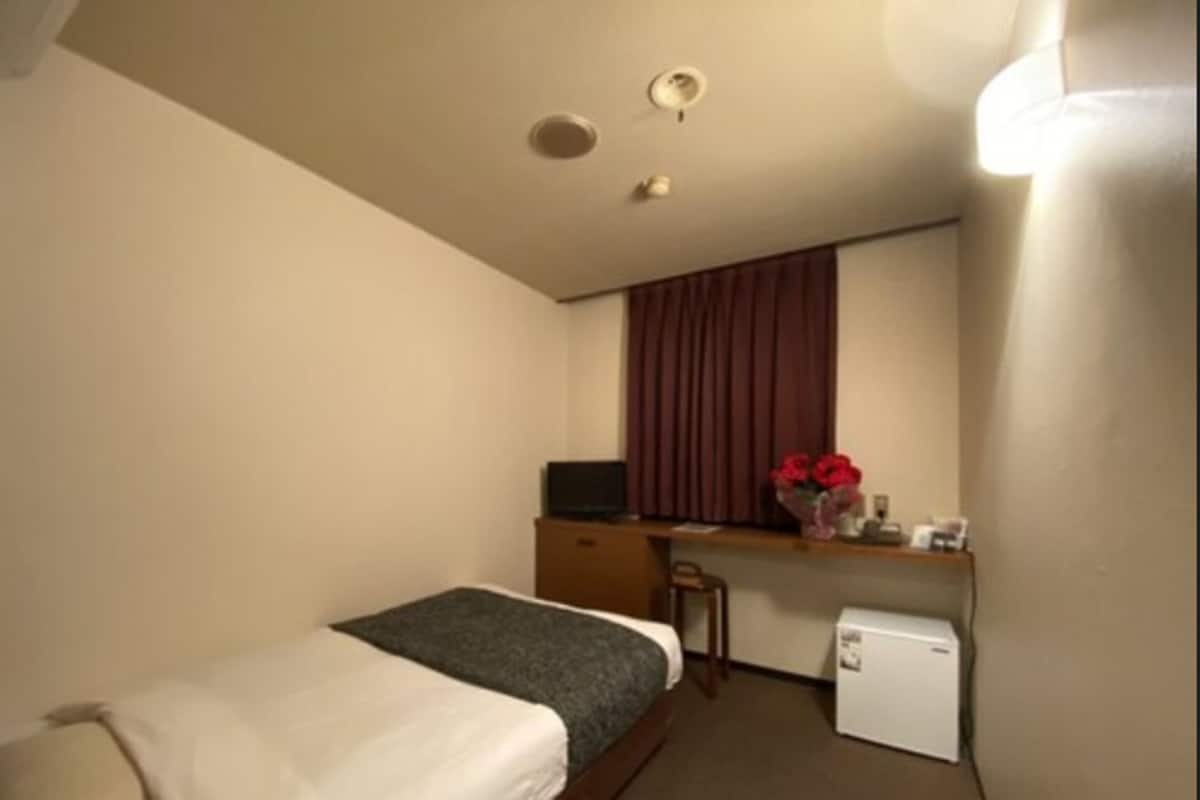 A small hotel room with a single bed, a desk with a computer, a small refrigerator, and a window with curtains.