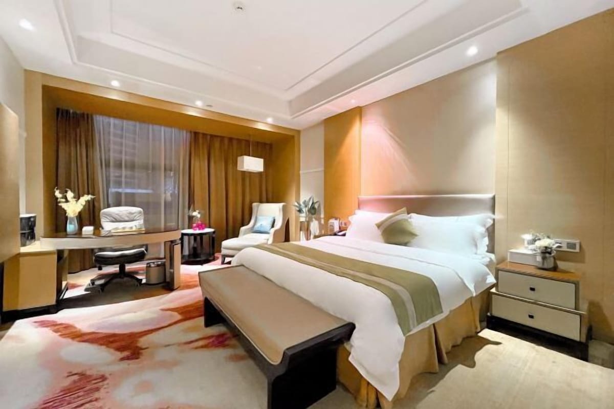 A hotel room with a large bed, a desk, a chair, a bench, and a nightstand with a lamp.
