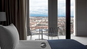 Habitación de hotel con cama, una mesita pequeña, dos sillas y un ventanal que ofrece vistas a la ciudad.