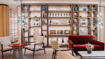 A modern living room with a red sofa, beige armchairs, and a coffee table. There is a bookshelf filled with books, wine bottles, and framed pictures.