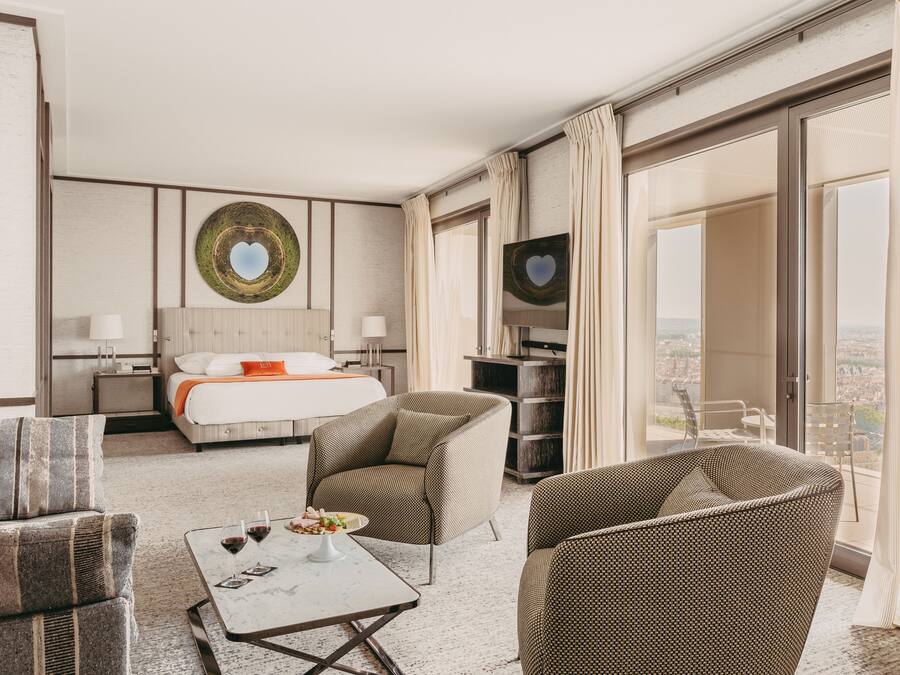 Une chambre d’hôtel comprenant un lit, deux fauteuils, une table basse, un téléviseur et une grande fenêtre avec des rideaux.