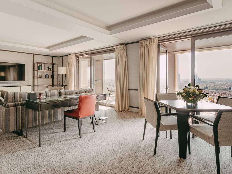 A modern hotel room with a sofa, dining table, and chairs, a TV, and a city view.