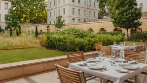 Un área de comedor al aire libre con mesas dispuestas para una comida, rodeada de follaje y un edificio grande al fondo.