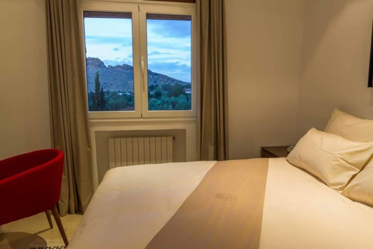 Une chambre d’hôtel avec un lit, une chaise rouge, une fenêtre donnant sur la montagne et un radiateur.